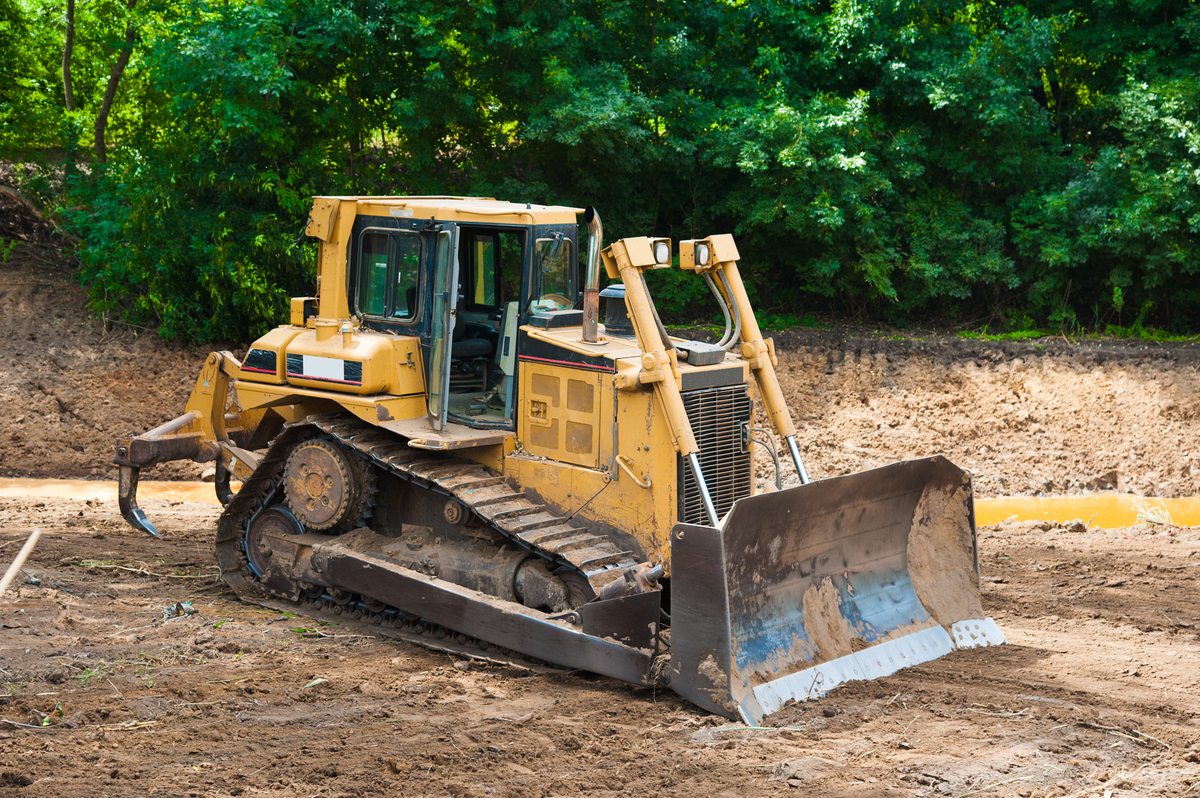 Yellow bulldozer parked on a dirt construction site near a wooded area, with muddy tracks and a large front blade visible